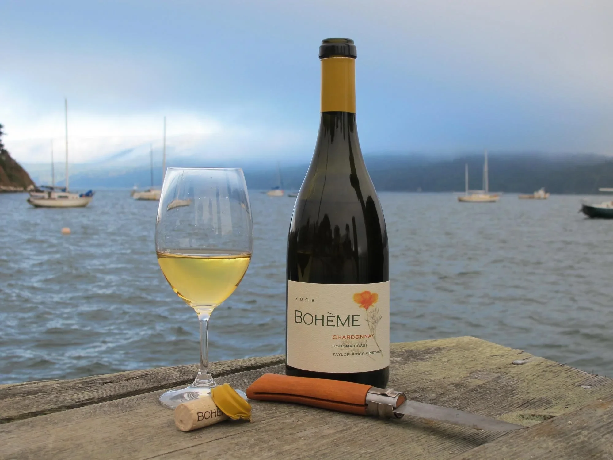 A bottle of Boheme Chardonnay wine, a wine glass filled with white wine, a corkscrew, and a cork with a yellow foil lie on a wooden table by the water. There are boats on the water with mountains and a cloudy sky in the background.
