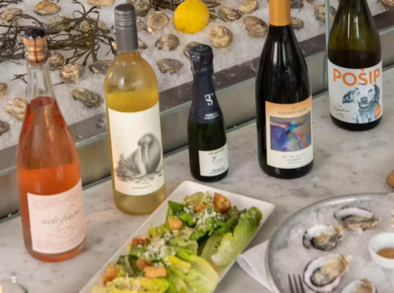 Various bottles of wine and a plate with oysters and salad on a marble countertop.