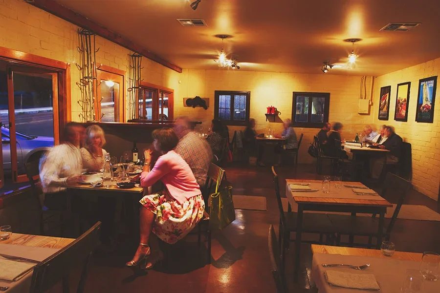 Interior of a warmly lit restaurant with wooden tables and chairs, some occupied by groups of people dining and conversing. The walls are decorated with framed art, and large windows with curtains overlook the street outside. Soft lighting creates a cozy ambiance.