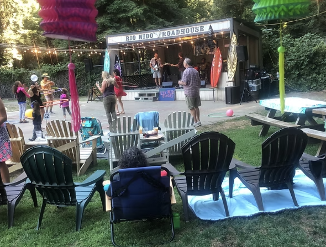 People enjoying an outdoor family gathering at a venue named 'Rio Nido Roadhouse A,' with a stage where a band is performing and children playing with hula hoops and balloons, surrounded by trees and string lights.