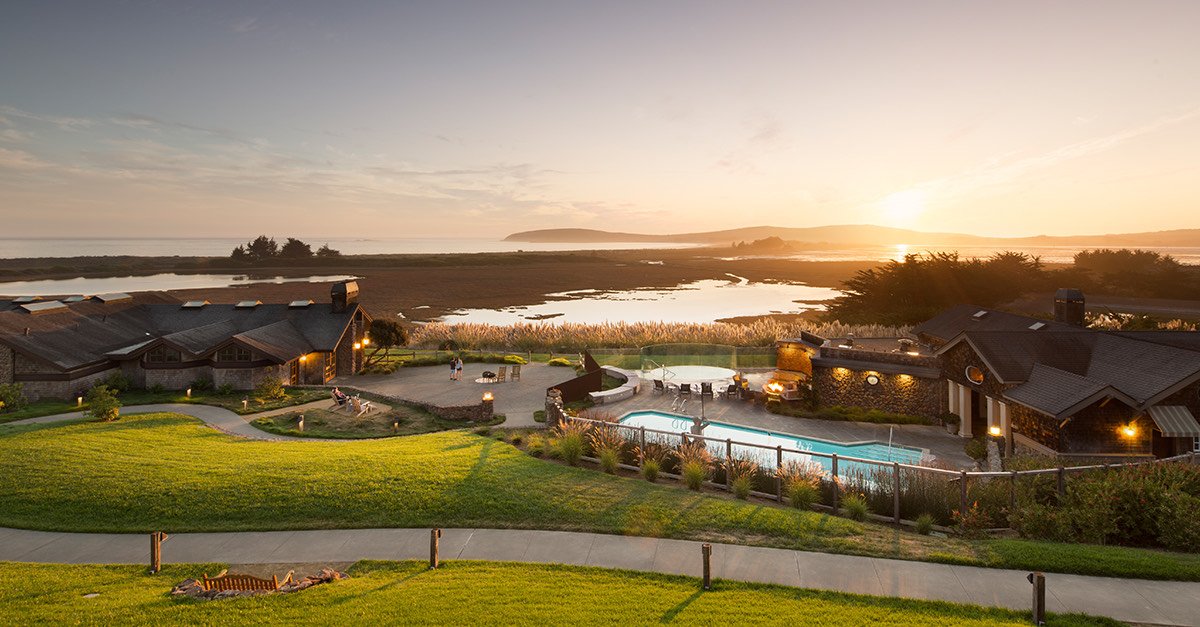 A scenic view of a luxurious resort with a swimming pool, surrounding buildings, and a body of water with an island in the background at sunset.