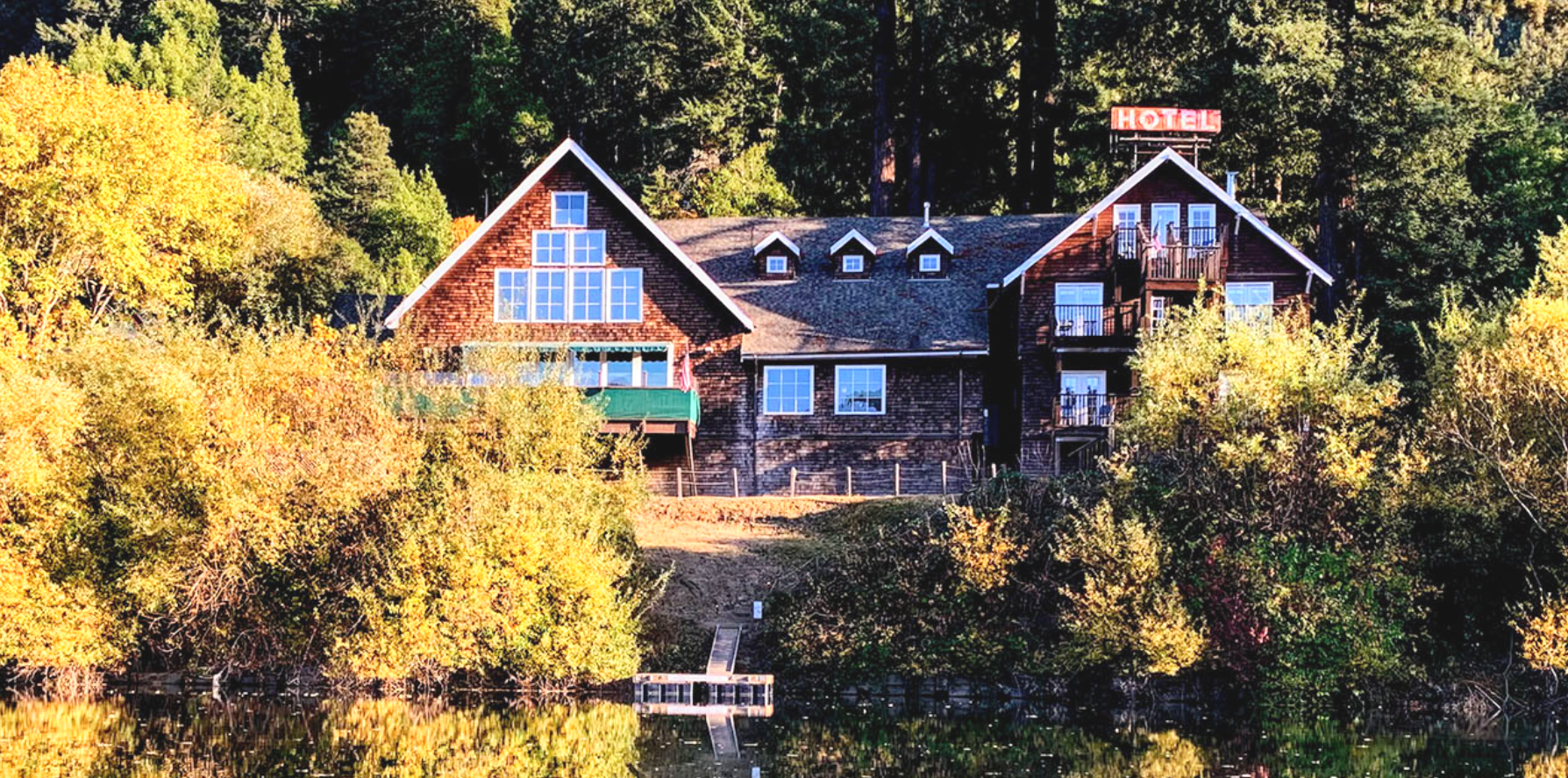 A rustic hotel with a brown wooden exterior, balconies, and a sign on the roof that says 'HOTEL', situated on a hillside surrounded by trees with autumn foliage, next to a body of water reflecting the scenery.