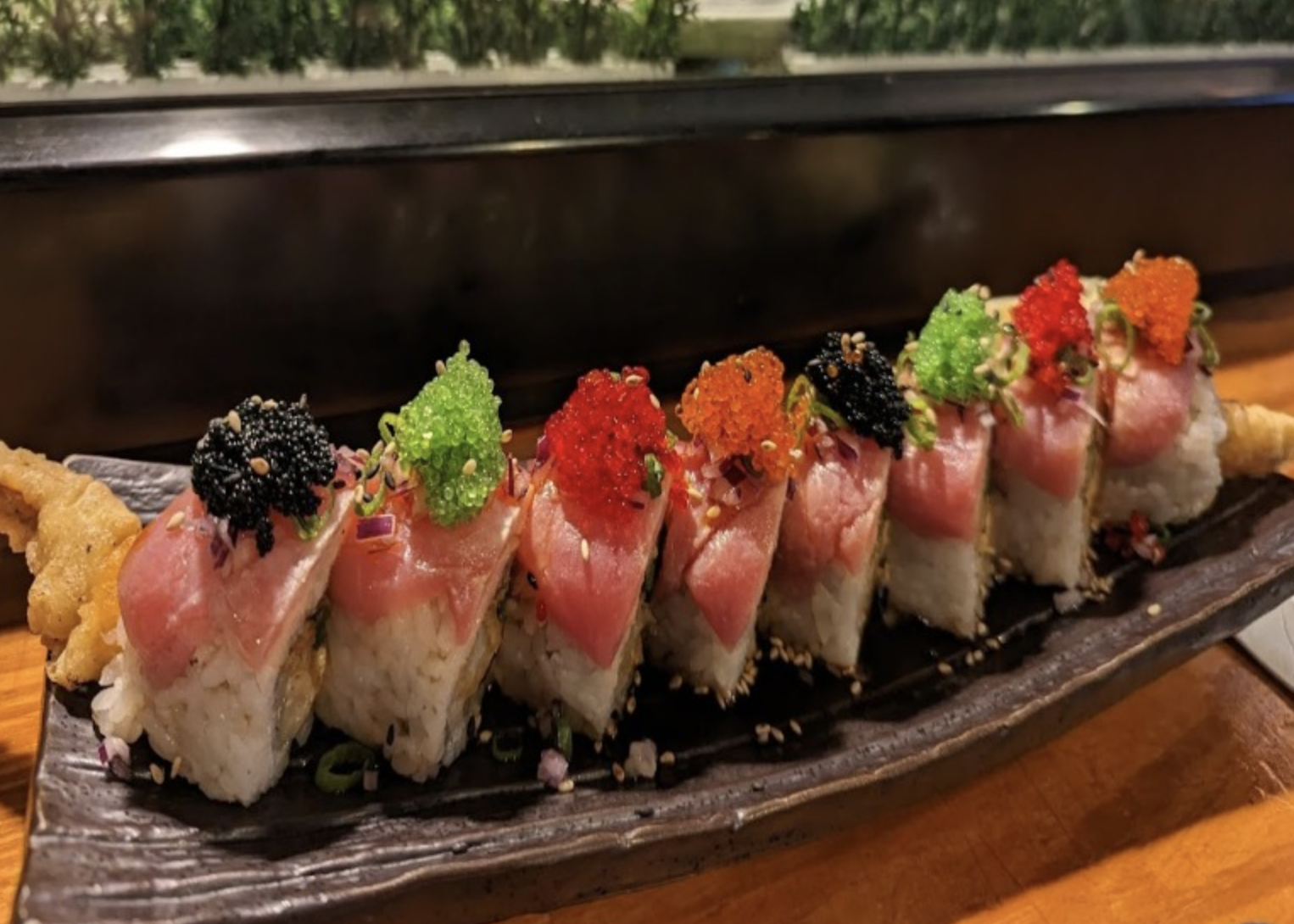 Sushi rolls topped with various colorful fish and roe, garnished with sesame seeds on a black ceramic plate.