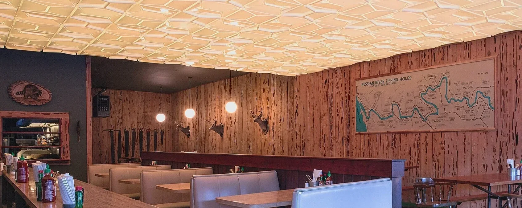 Interior of a rustic restaurant with wood-paneled walls, deer head mounts, and large map on the wall showing Russian River fishing holes. Booths and tables are set with condiments and utensils. Hanging lights illuminate the space.