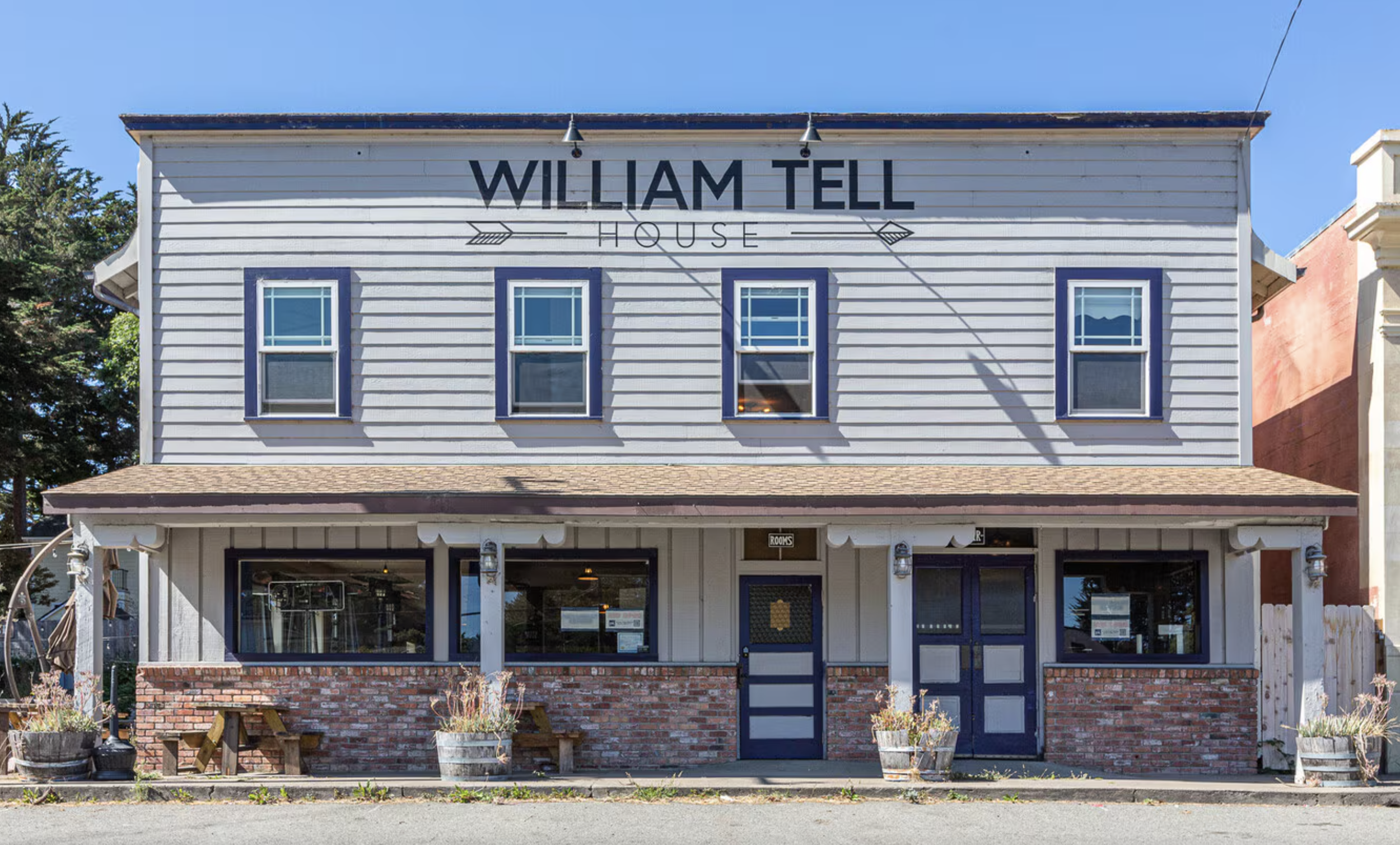Front view of William Tell House, a two-story building with gray siding and blue window frames, brick lower facade, and a sign with its name and an arrow symbol on top.