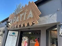 Storefront with wooden sloped sign reading 'TAPATIA' and a black and white logo above the entrance.