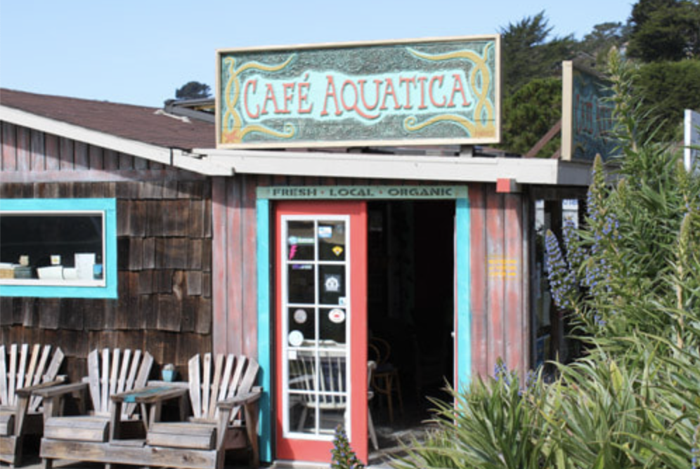 Exterior of Café Aquatica with a colorful sign, wooden siding, outdoor seating, and lush greenery.