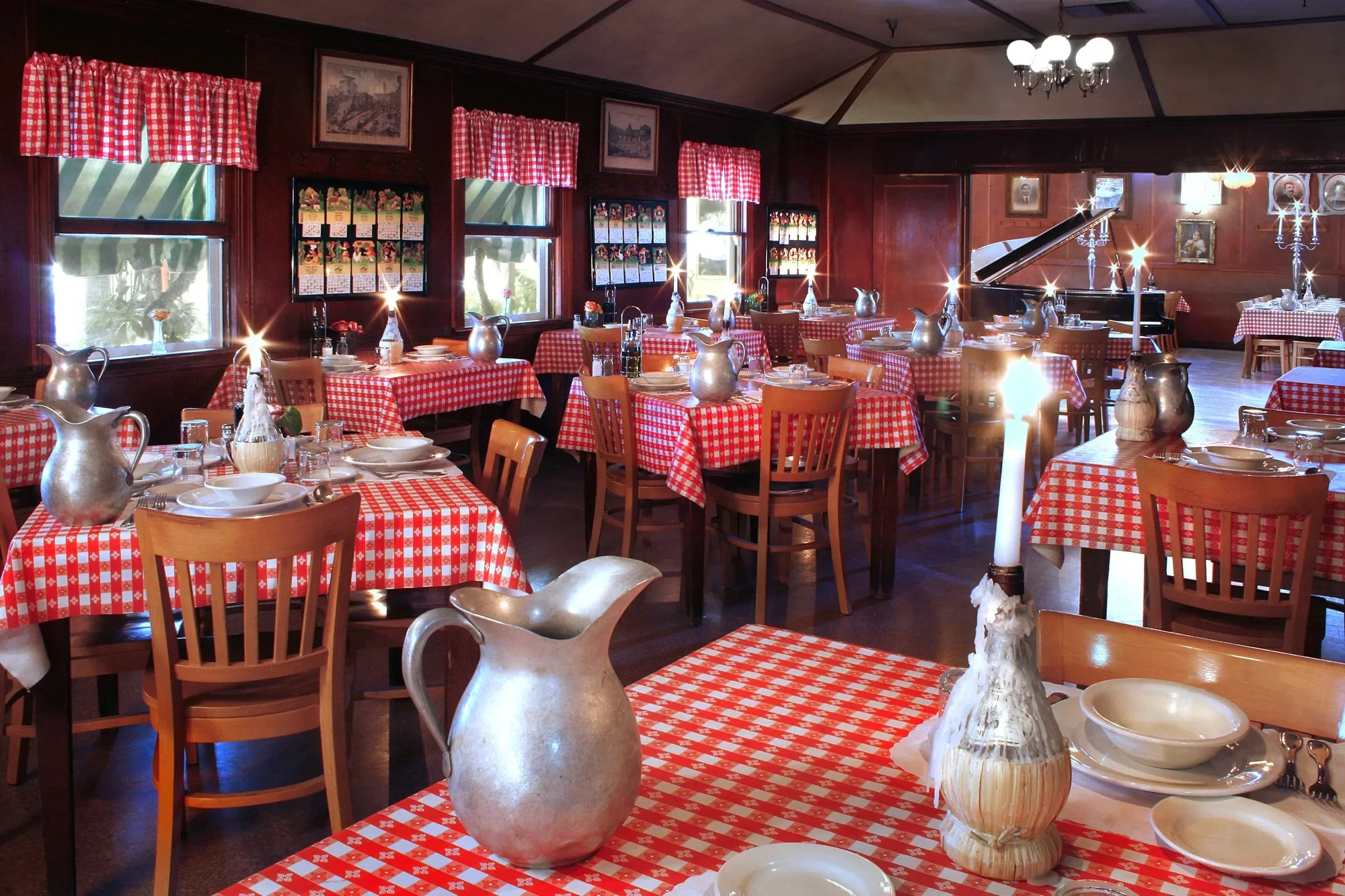 A cozy restaurant with red and white checkered tablecloths, wooden chairs, and silver pitchers on the tables. The walls are adorned with framed pictures and small decorative windows with matching checkered valances. There is a grand piano in the background and lit candles on the tables, creating a warm atmosphere.