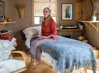 An elderly woman sitting on a bed in a cozy, well-lit bedroom with wooden shelves, a window with blinds, and decorative items.