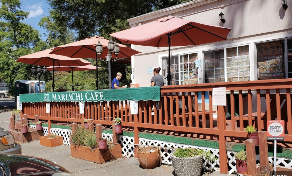 The outdoor patio of El Mariachi Cafe features brown wooden railings, pink umbrellas, and potted plants. Two people are standing at the counter, and the cafe has large windows with colorful signs inside.
