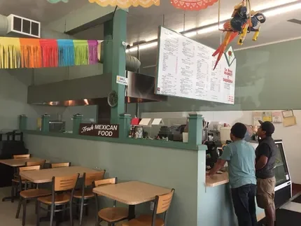 Inside a Mexican restaurant with colorful decorations, a menu board hanging above the counter, and two employees working behind the counter.