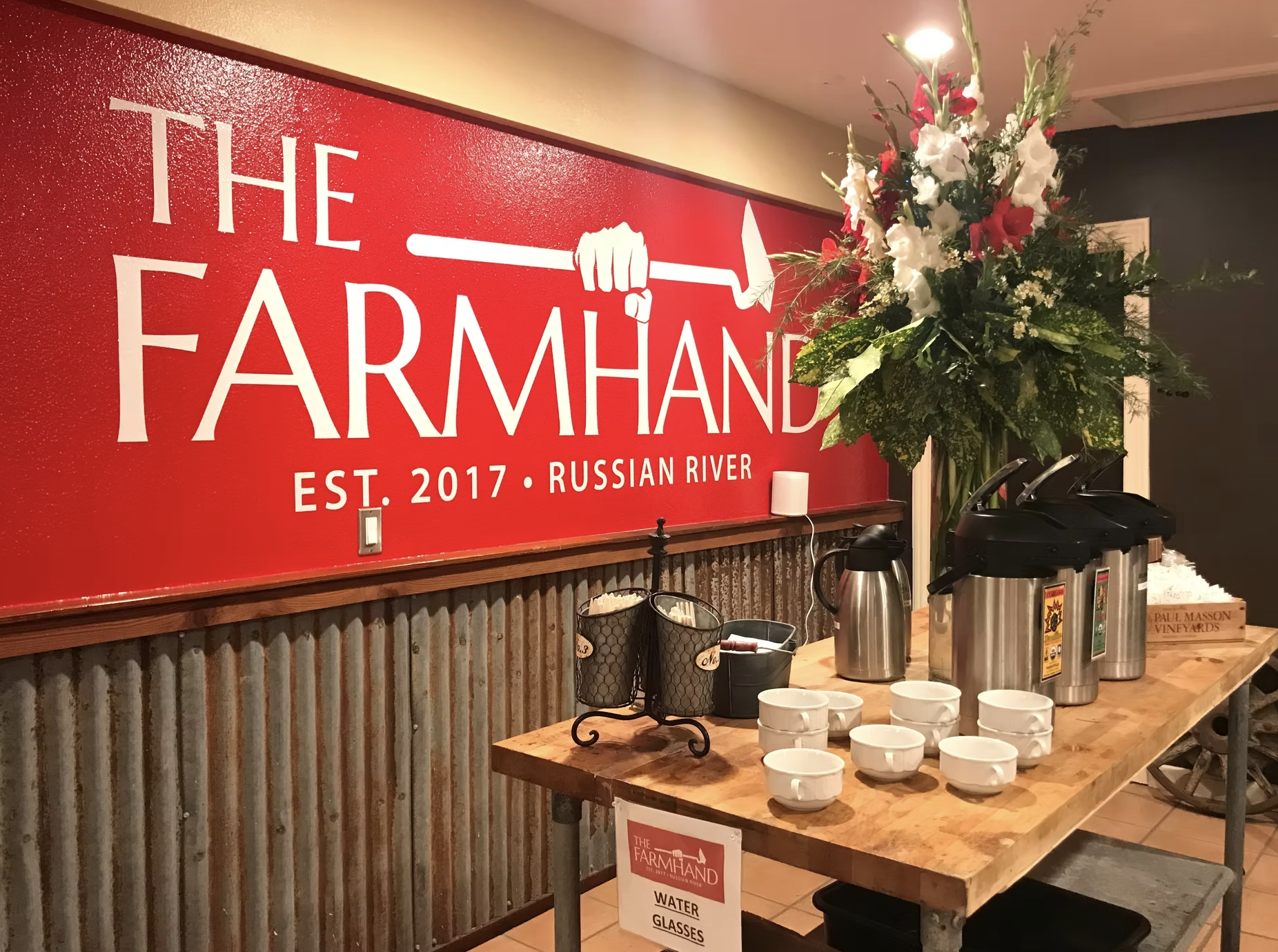A table with water glasses and coffee dispensers in front of a large floral arrangement. A sign on the table reads 'Water Glasses' and a large red sign on the wall says 'The Farmhand EST. 2017, Russian River'.