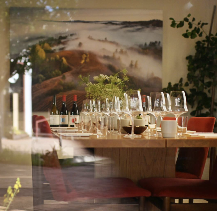 A dining table set with wine bottles, glasses, and tableware, with a landscape painting and a plant in the background, viewed through a window.