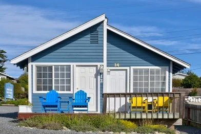 Blue house with yellow and blue outdoor furniture in front, wooden porch, white door, and large windows, under a blue sky.