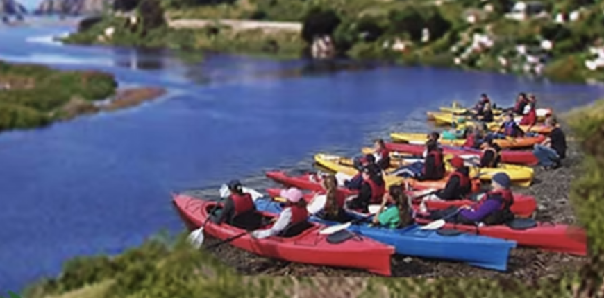 People preparing to kayak on a river with colorful kayaks and a scenic landscape in the background.