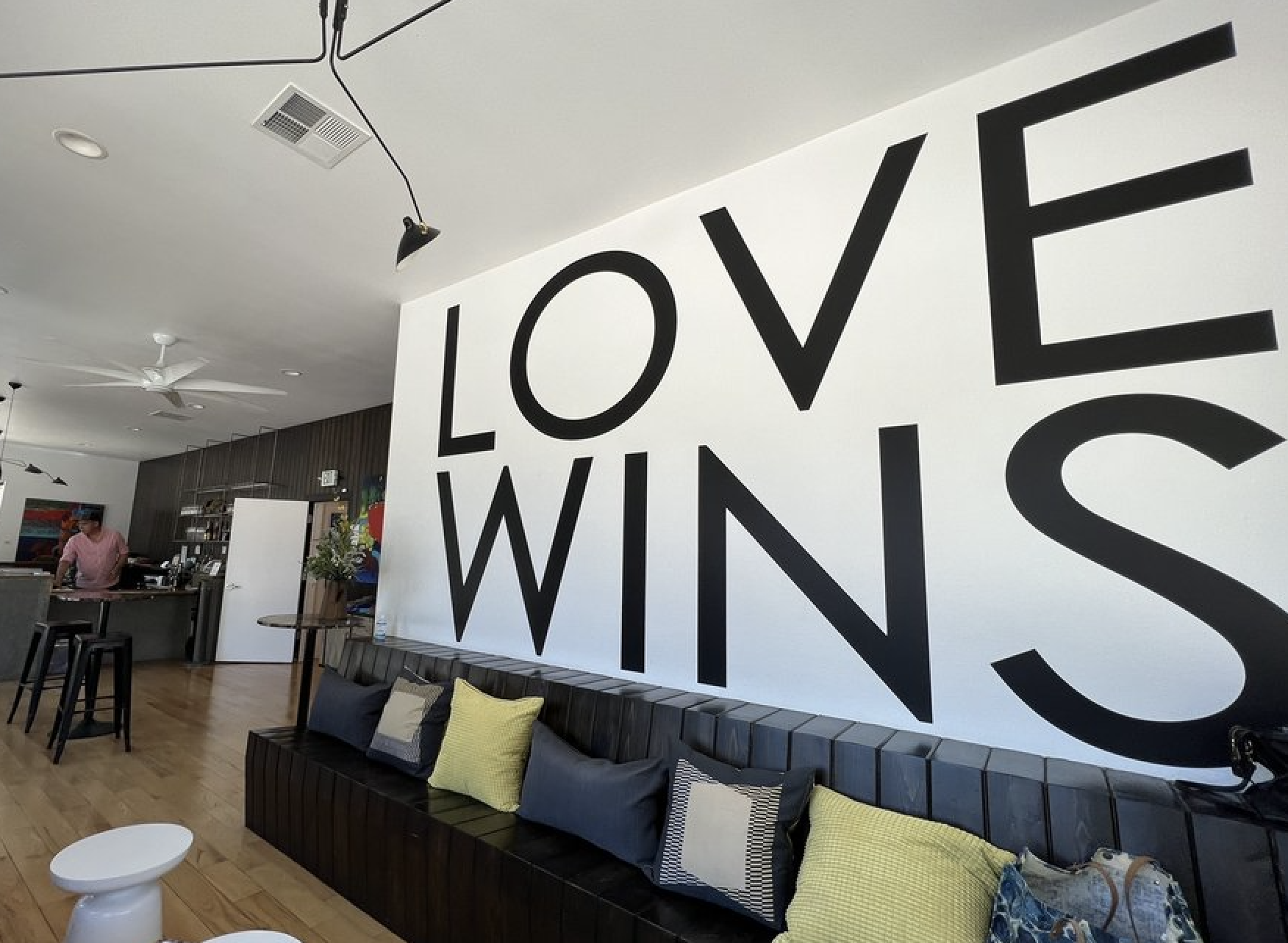 Interior of a modern, stylish space with a large white wall displaying the words "LOVE WINS" in bold black letters. There are black and yellow cushions on a black seating area, a small white side table, and a person working at a counter in the background. The space has a wooden floor, ceiling fans, and contemporary decor.