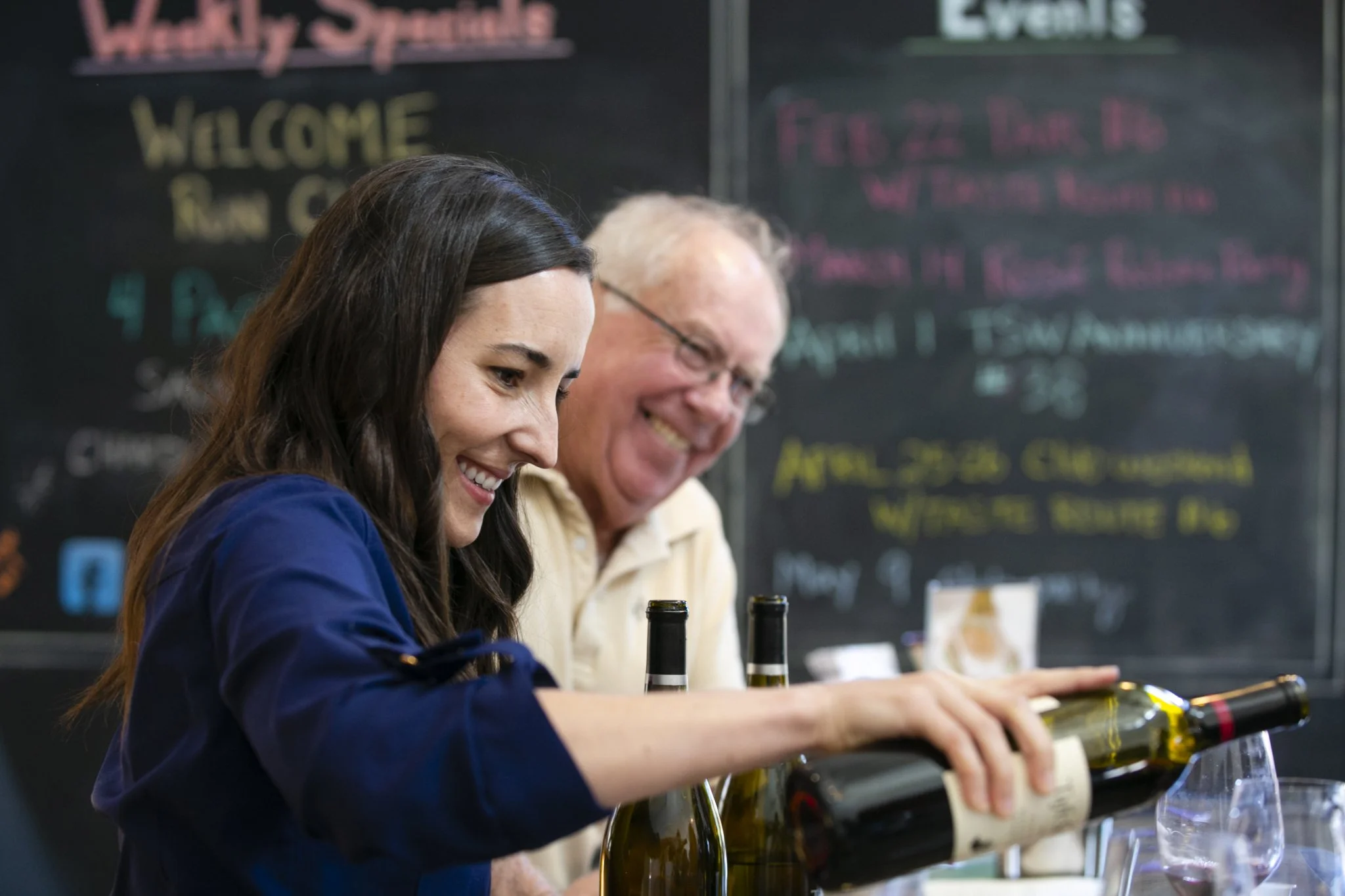 Woman and man pouring wine at a bar or restaurant, smiling and enjoying conversation, with a blackboard menu in the background.