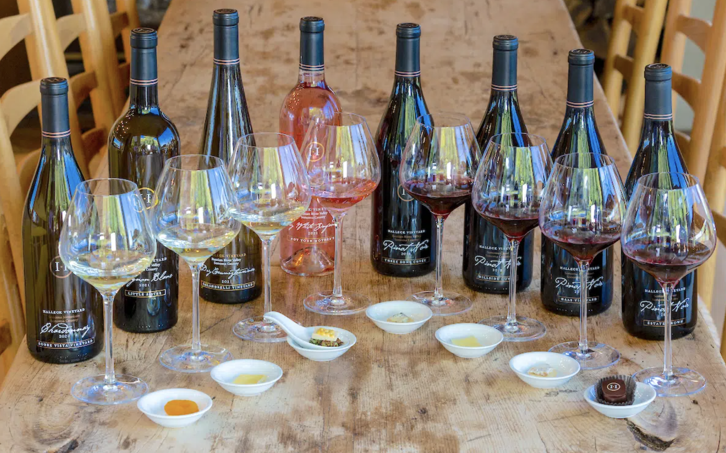 Assorted bottles of wine and five wine glasses filled with white, rosé, and red wine on a wooden table, with small dishes of tasting notes and chocolates