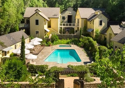 Aerial view of a residential complex with a swimming pool, surrounded by multiple yellow and gray houses with balconies and lush green trees.