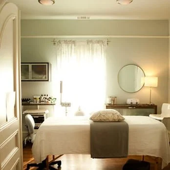 A bedroom with a bed, window with curtains, round mirror, desk, and chair.