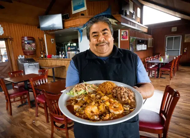 A man smiling and holding a large plate of food inside a restaurant with wooden walls and red chairs.