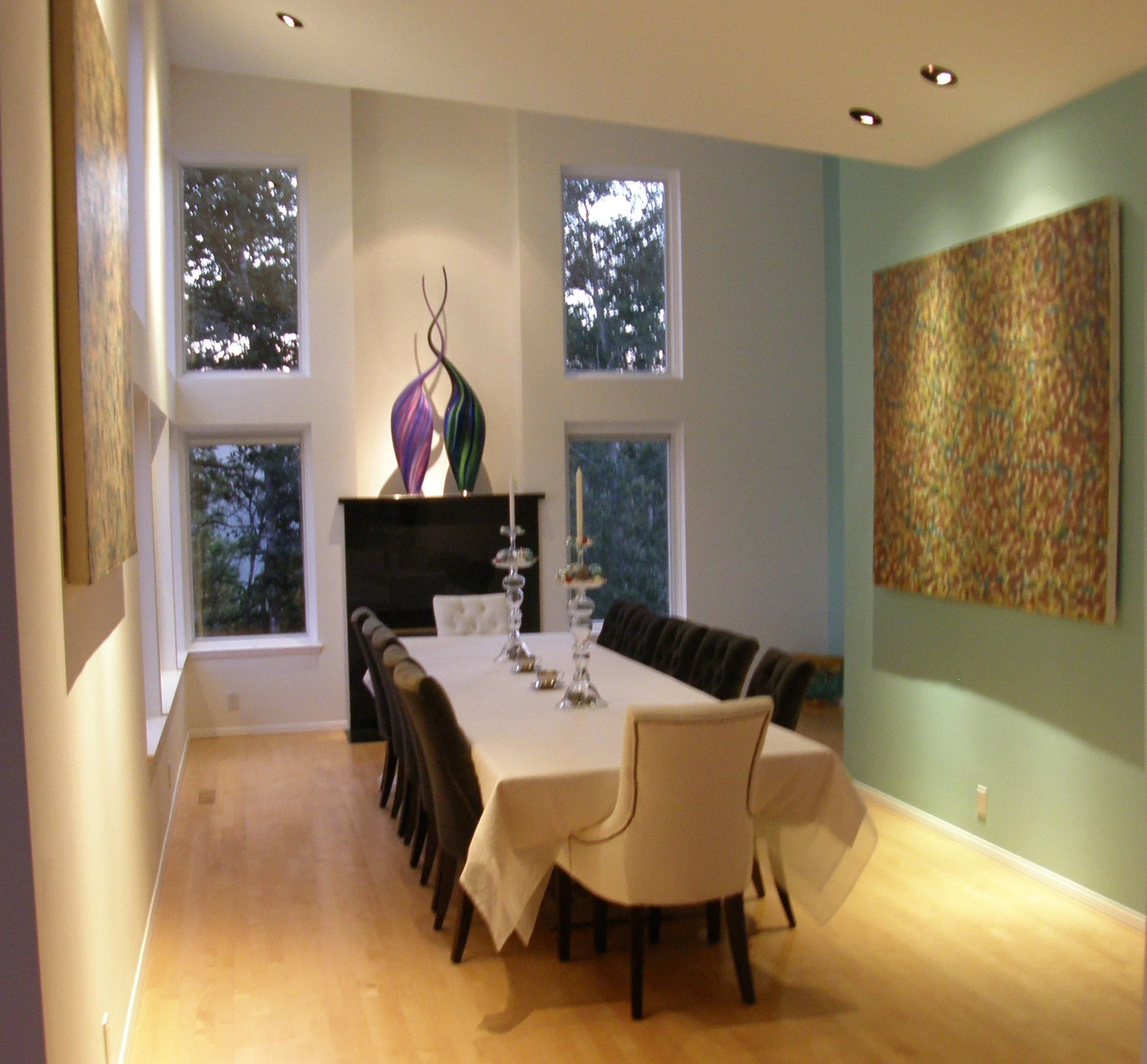 A dining room with a long table covered with a white tablecloth, surrounded by multiple chairs, including one with cream-colored upholstery. The room has light-colored hardwood flooring, several windows letting in natural light, a black fireplace with colorful glass sculptures on top, and decorative candle holders on the table. Artwork is displayed on the walls.