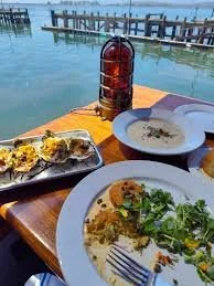 Seafood meal on a wooden table by the water with a grilled fish, a bowl of clam soup, and a plate with sauce and herbs, near a dock.