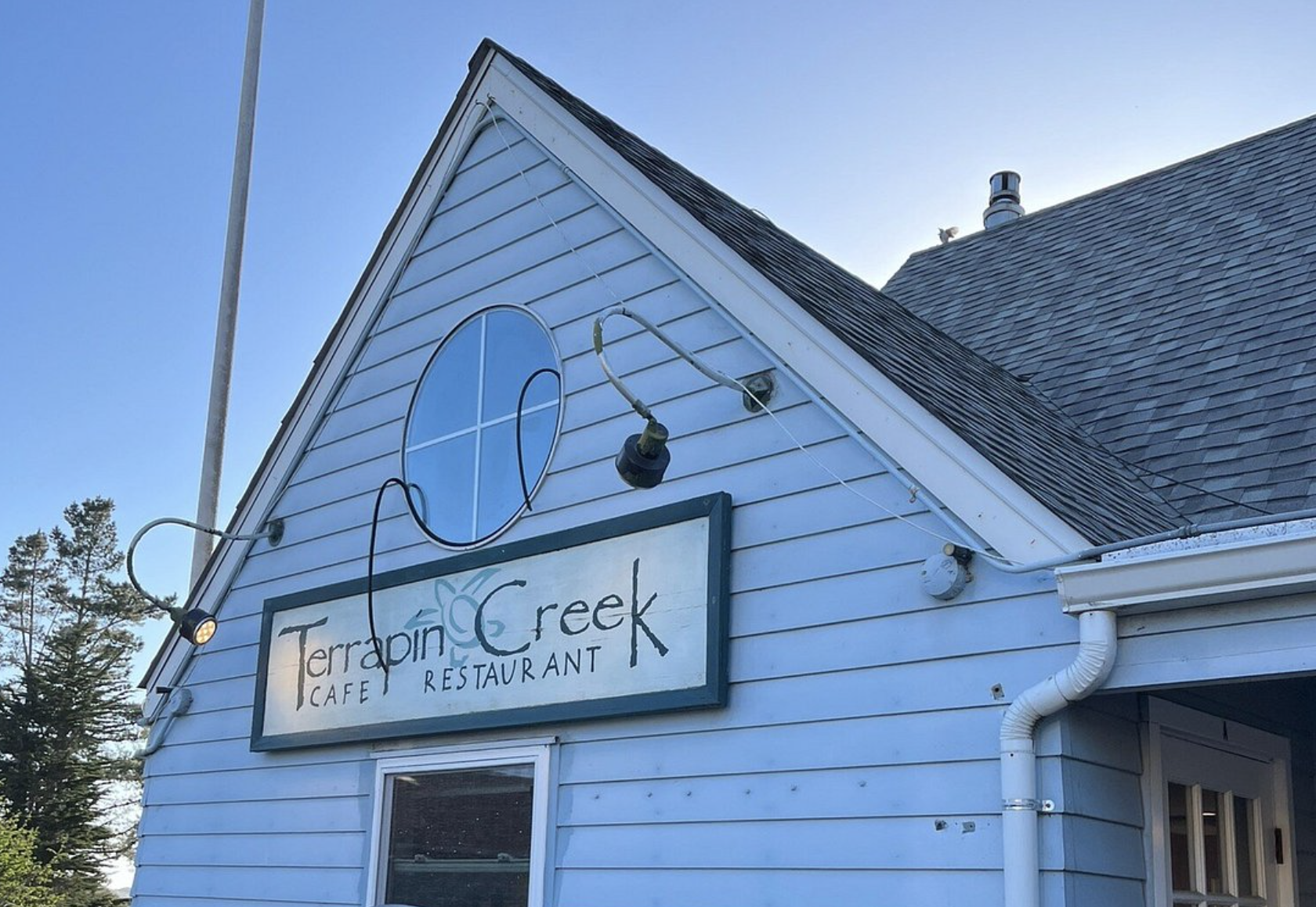 Exterior of a blue house with a sign that reads 'Terrapin Creek Café Restaurant.' A light is turned on above the sign, and a small window is visible below. There are trees in the background and a blue sky.