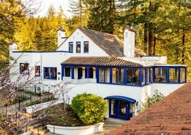 A large white house with multiple sections, blue accents, and roof chimney, surrounded by trees and some landscaping, under a clear sky.