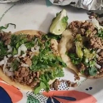 Two tacos filled with seasoned ground beef, chopped onions, cilantro, and diced green peppers on a plate with a lime wedge.