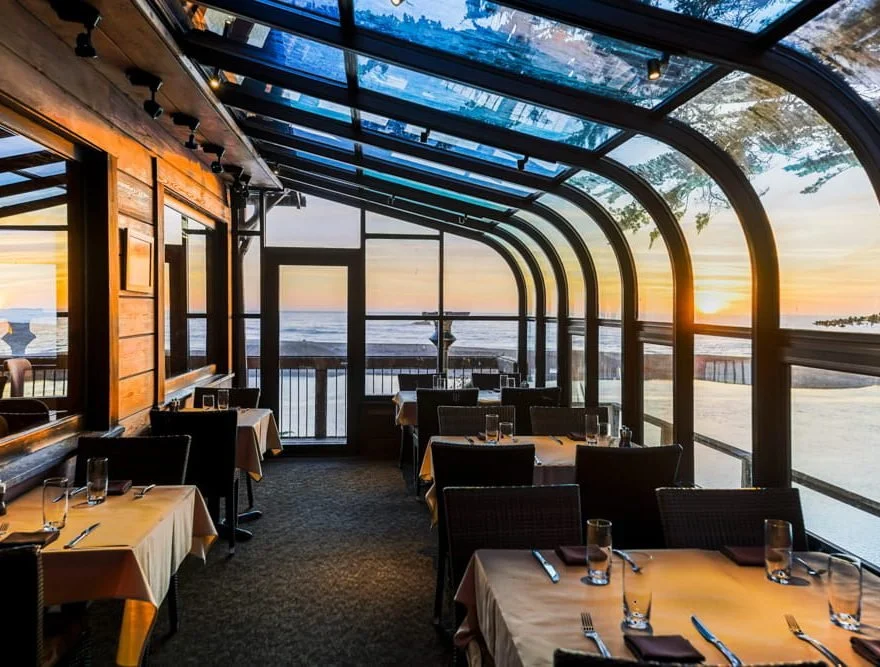 Inside a restaurant with large glass windows overlooking the ocean at sunset, dining tables set with glasses, silverware, and napkins.