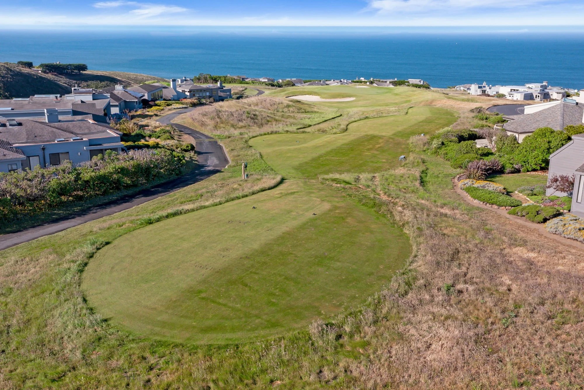 Aerial view of a coastal golf course with green fairways, sand bunkers, residential houses, and the ocean in the background.