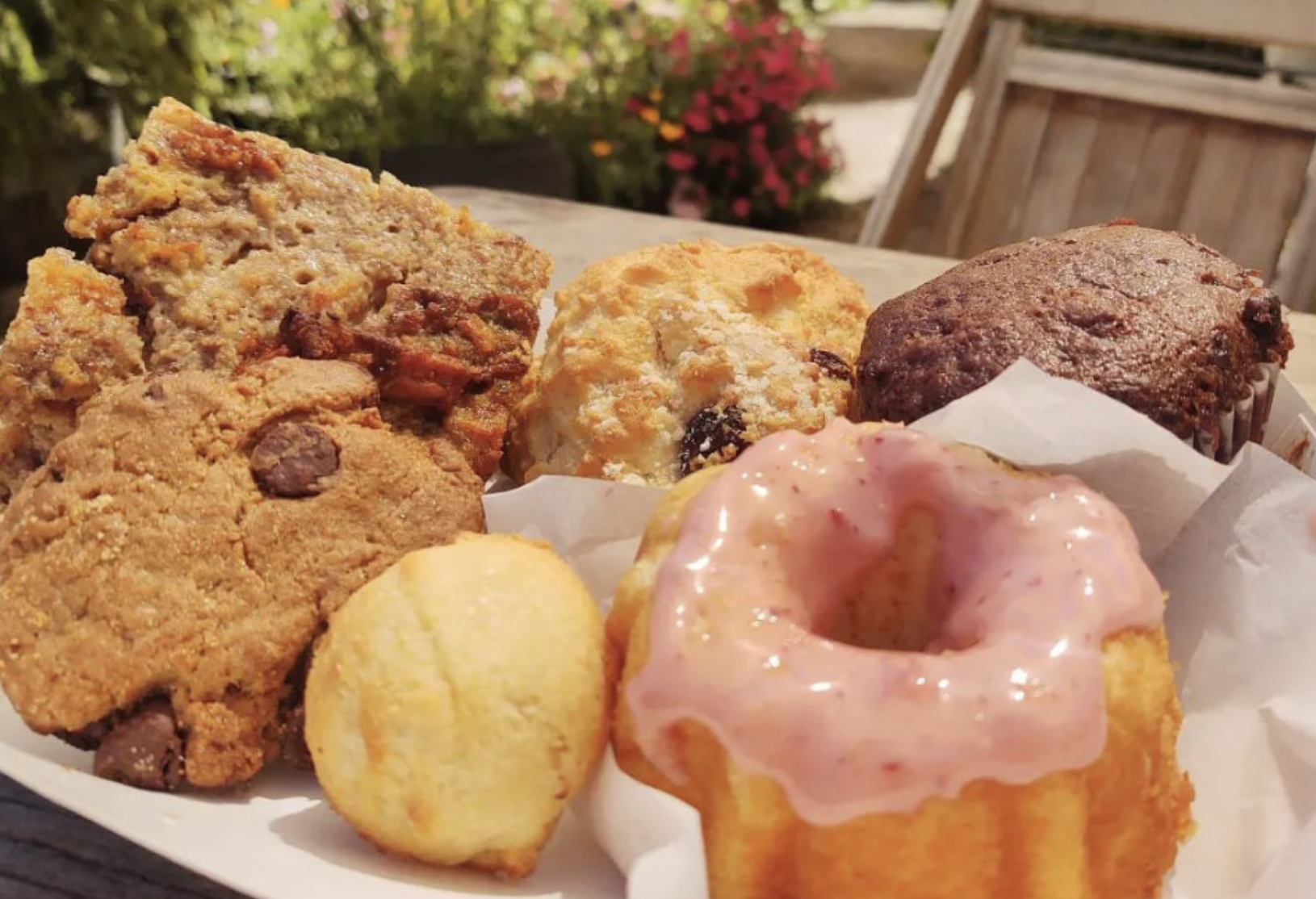 Assorted baked goods including cookies, muffins, and a donut with pink glaze, arranged on a tray outdoors.