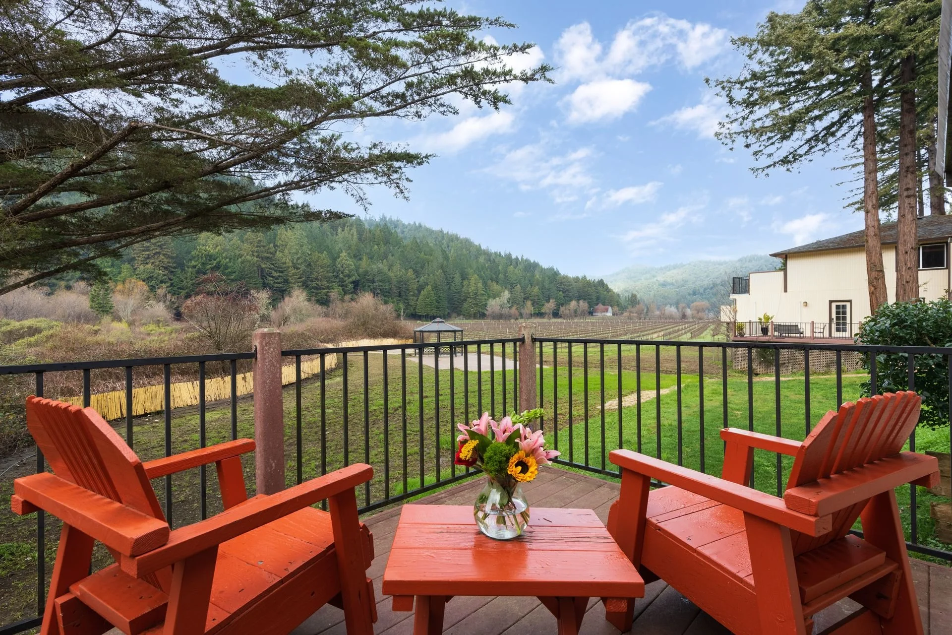 Two orange Adirondack chairs facing a small orange table with a glass vase of pink lilies, yellow sunflowers, and greenery, on a deck overlooking a green lawn, trees, and distant mountains under a partly cloudy sky.