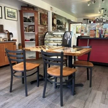 Interior of a café with wooden tables and black chairs, a display case, and shelves with various items, with a counter in the background.