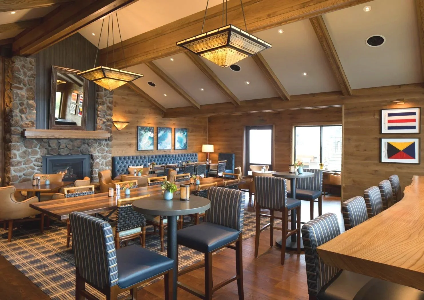 A cozy restaurant interior with wooden walls and ceiling, decorated with framed artwork and a stone fireplace. The space features tables and chairs, some with striped upholstery, arranged for dining, with a large window letting in natural light.