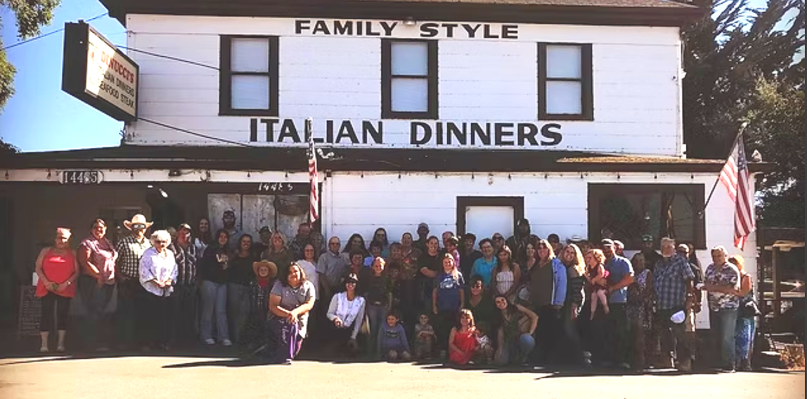 Group of people standing in front of a building with signs advertising Italian dinners and family-style meals, with American flags on either side.