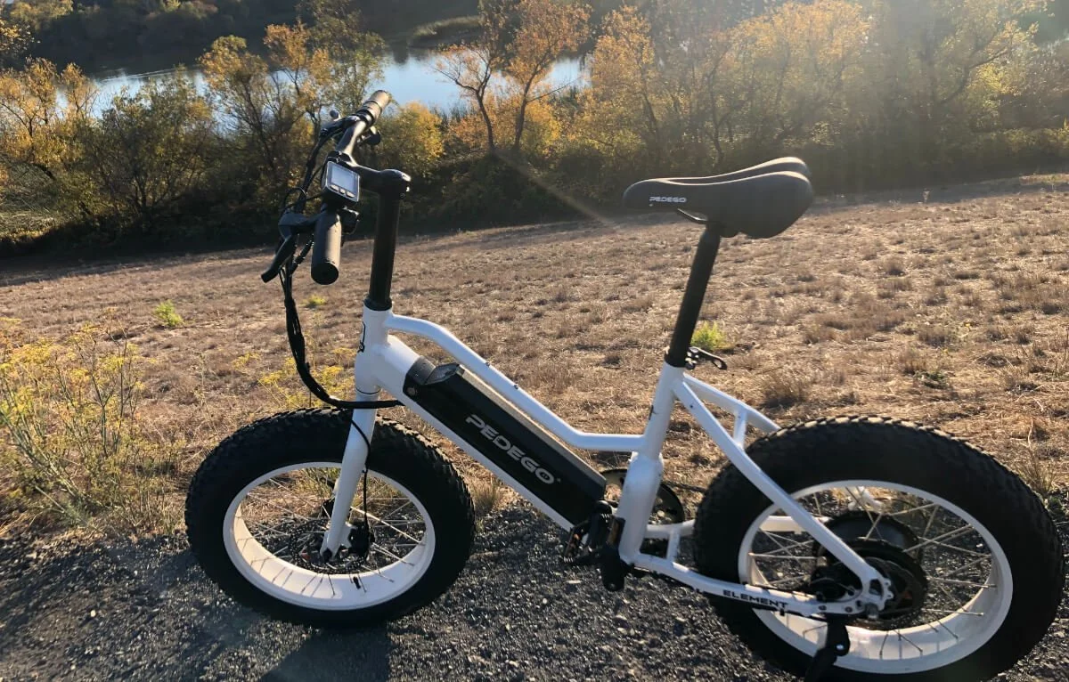 White electric mountain bike with fat tires parked on a dirt path near a tree-lined body of water during sunset.