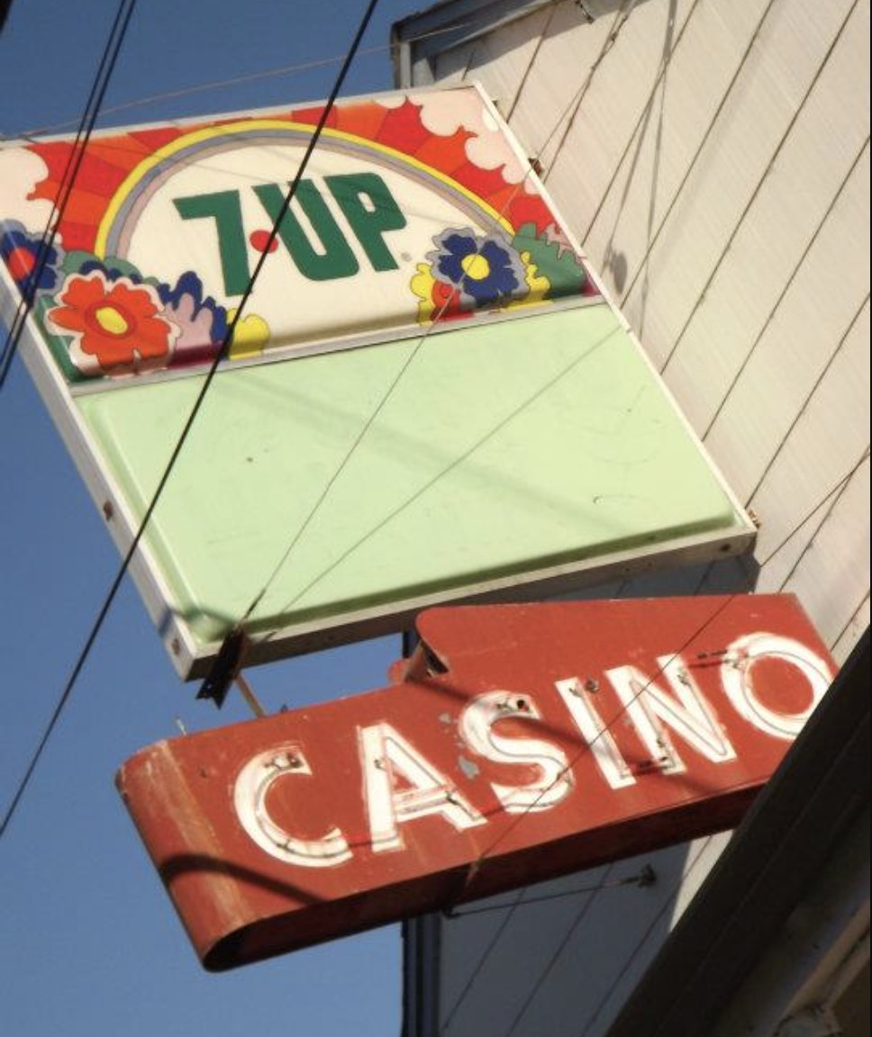 Vintage neon Casino sign hanging above a building, with a 7UP advertisement sign featuring colorful flowers, against a clear blue sky.