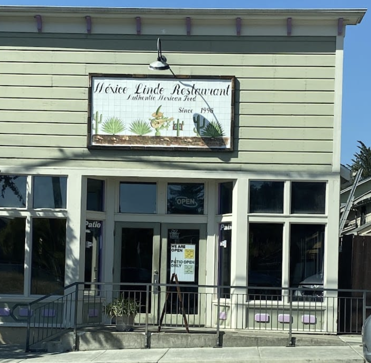 Exterior view of Mexice Linde Restaurant with a sign above the entrance, featuring illustrated cacti and a sombrero, and an open sign in the window.