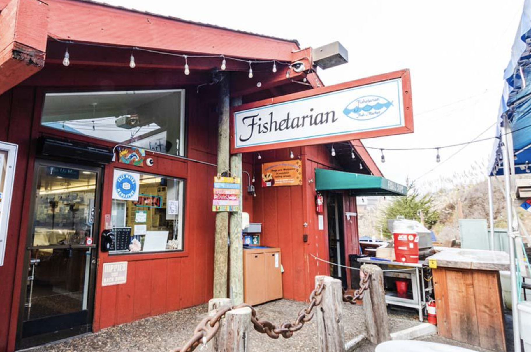 Exterior of a red wooden building with a sign that reads 'Fishetarian.' The building has string lights hanging above and a window with a small sign that says 'blue fish.' There is a wooden post with a chain barrier in front of the entrance, and outdoor equipment including a red cooler and a fire extinguisher are visible.