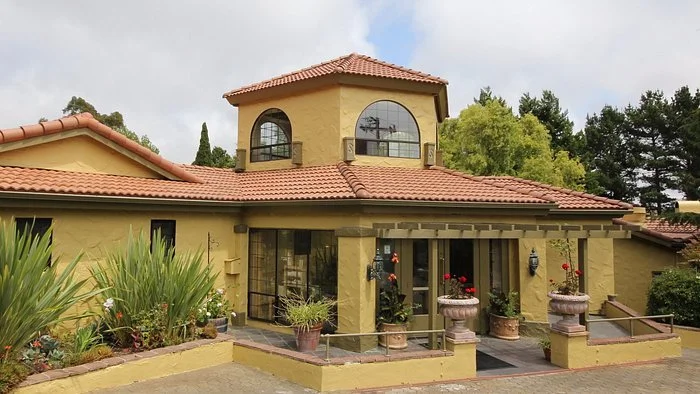 A yellow stucco house with a red tile roof, featuring arched windows and a second-story tower, surrounded by lush green trees and plants.