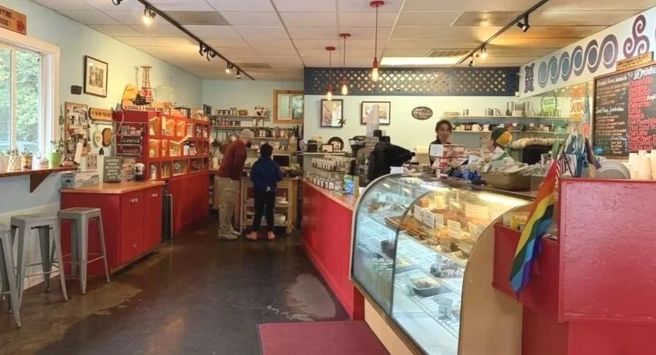 Interior of a cozy cafe with a counter display of baked goods, red counters, a rainbow Pride flag, and customers ordering.