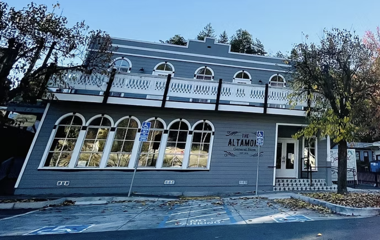 A two-story grey building with white trim and arched windows, labeled 'The Altamont General Store.' There's a balcony on the second floor and a parking lot in front with designated handicap parking spaces.