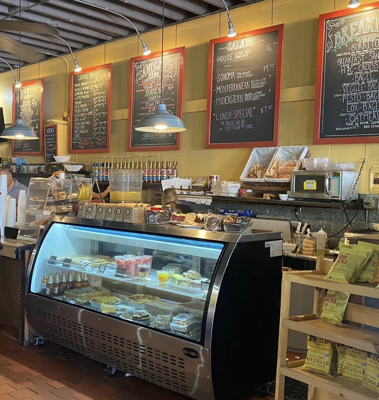 Inside a bakery or cafe with a display case of desserts and pastries, chalkboard menus hanging on yellow walls, and various food items on the counter.