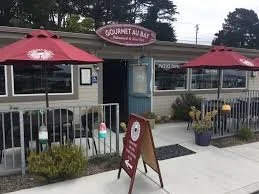 Exterior of a small diner or cafe with a sign that reads 'Gourmet Bay' and outdoor seating with red umbrellas.
