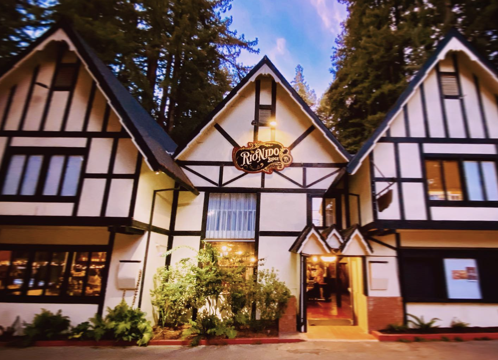 Exterior of a Tudor-style building with black wood beams and white walls, illuminated at dusk, with a sign that reads "Rio Linda Lodge" in the center, surrounded by trees and plants.