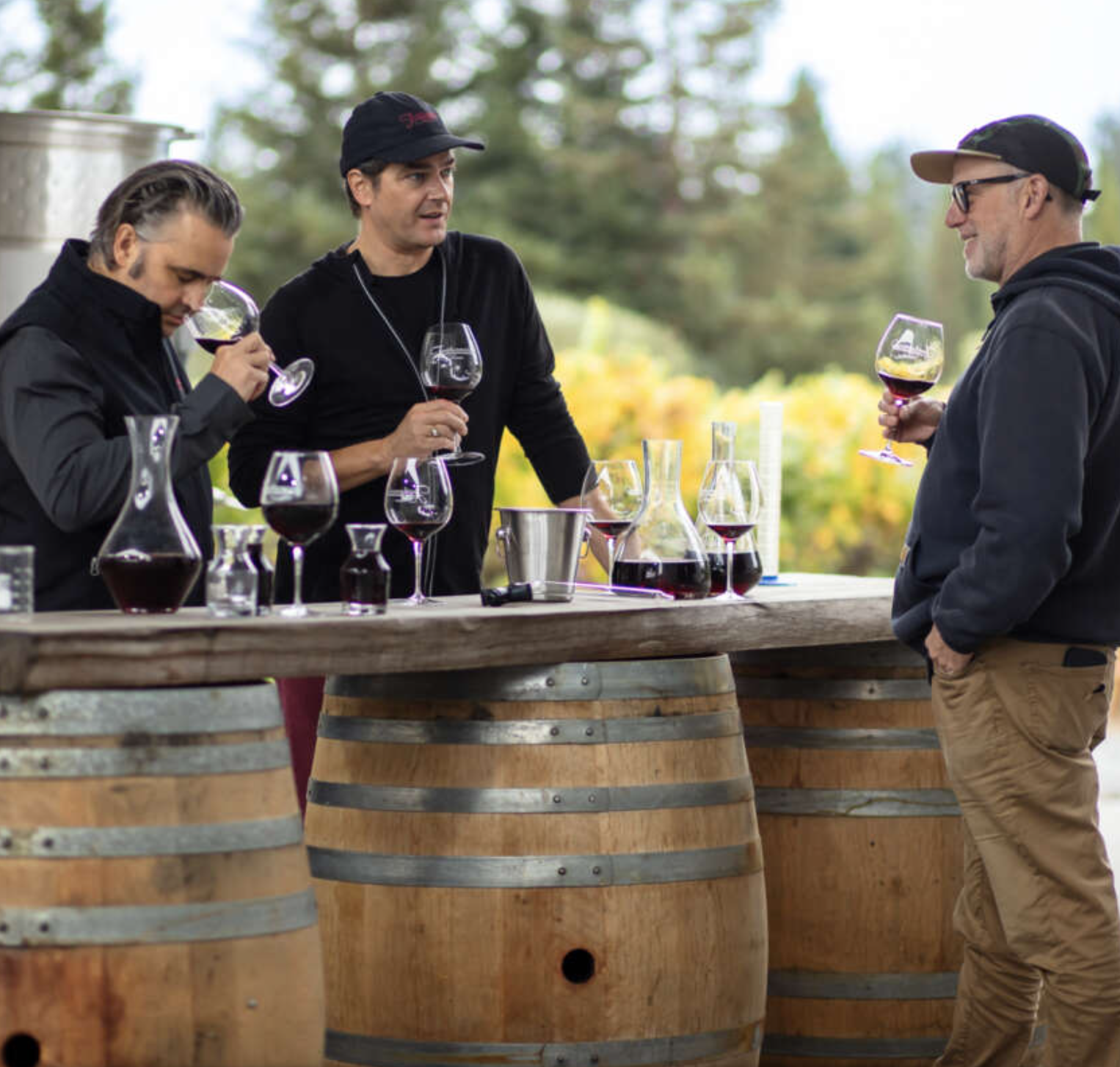 Three men toasting with glasses of red wine at an outdoor wine tasting event, with wine bottles and glasses on a table made of barrels, set against a backdrop of trees and yellow flowers.