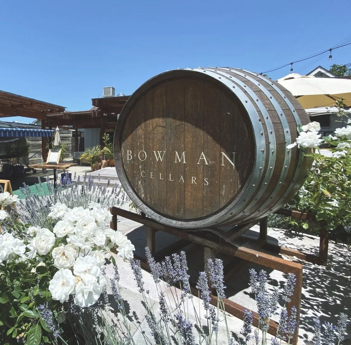 Wooden wine barrel with metal bands bearing the sign 'Bowman Cellars', surrounded by white and purple flowers at an outdoor winery or tasting room.