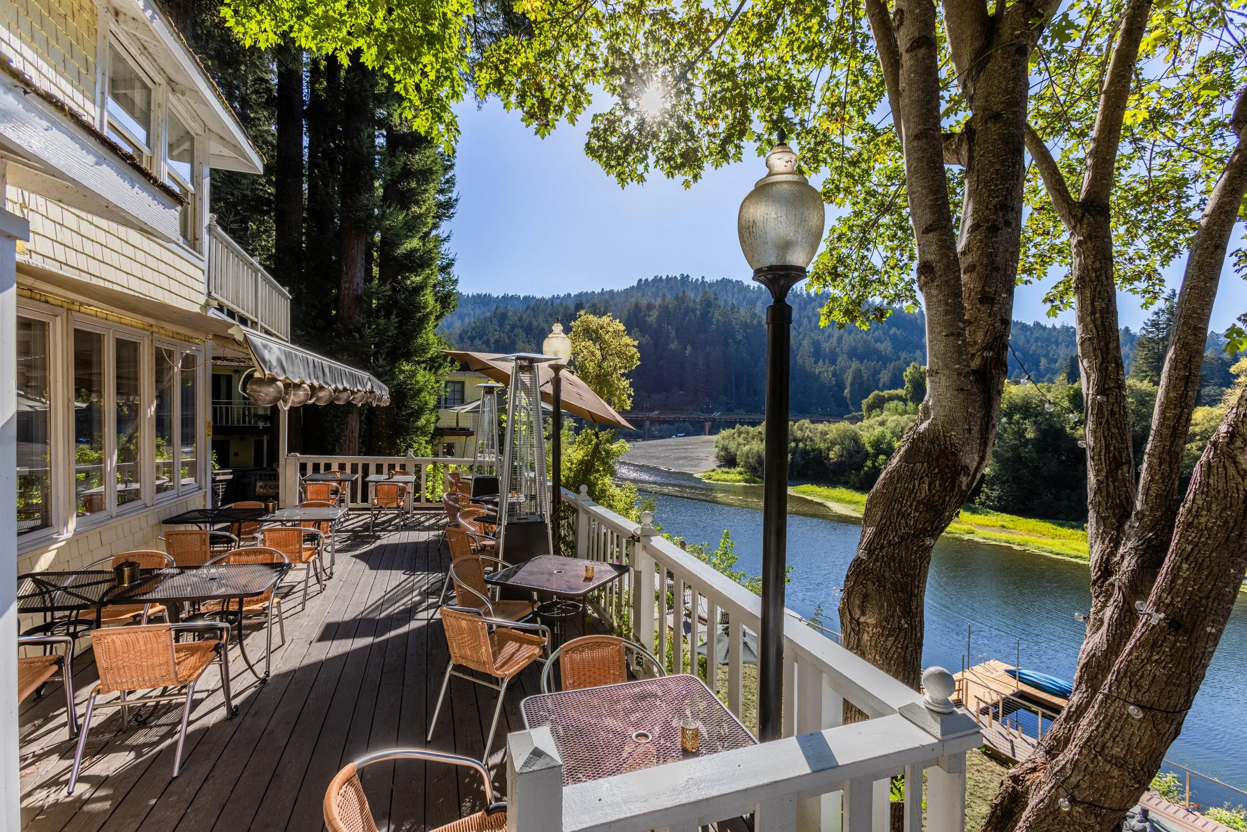 Empty outdoor restaurant patio with tables and chairs overlooking a river, surrounded by trees and hills on a sunny day.